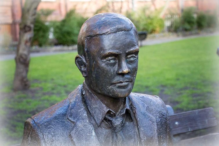 Eine Bronze-Skulptur – Alan Turing in ernster Miene – vor dem grünen Hintergrund des Sackville Parks in Manchester.