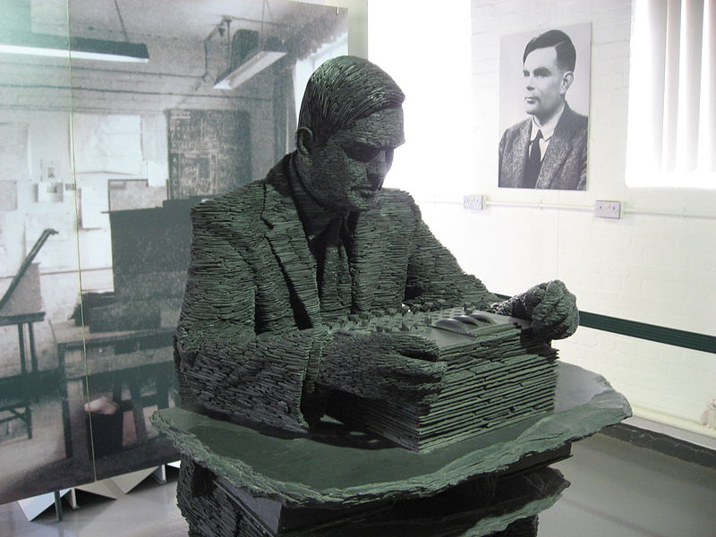 Eine grau-grüne Schiefer-Skulptur zeigt Alan Turing, streng geschitelt, sinnierend über einer ‚Enigma’-Codiermaschine im Aussehen einer Schreibmaschine.