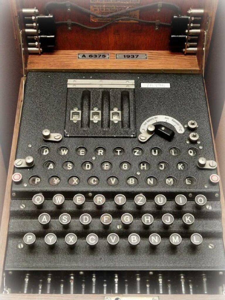 Die Chiffriermaschine Enigma im Aussehen einer Schreibmaschine im aufgeklappten Zustand. Typenhebel und ein Holzgehäuse verbergen das komplexe Getriebe aus ineinandergreifenden Walzen.
