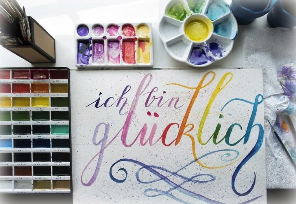 Schwungvoll, in Regenbogenfarben gestaltete Schriftzeile "Ich bin glücklich" im Handlettering-Stil, umgeben von Aquarell-Farbkasten und Palette.